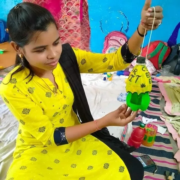 A young woman in a yellow dress holds a handcrafted hanging decoration made from a recycled plastic bottle in a colorful setting.