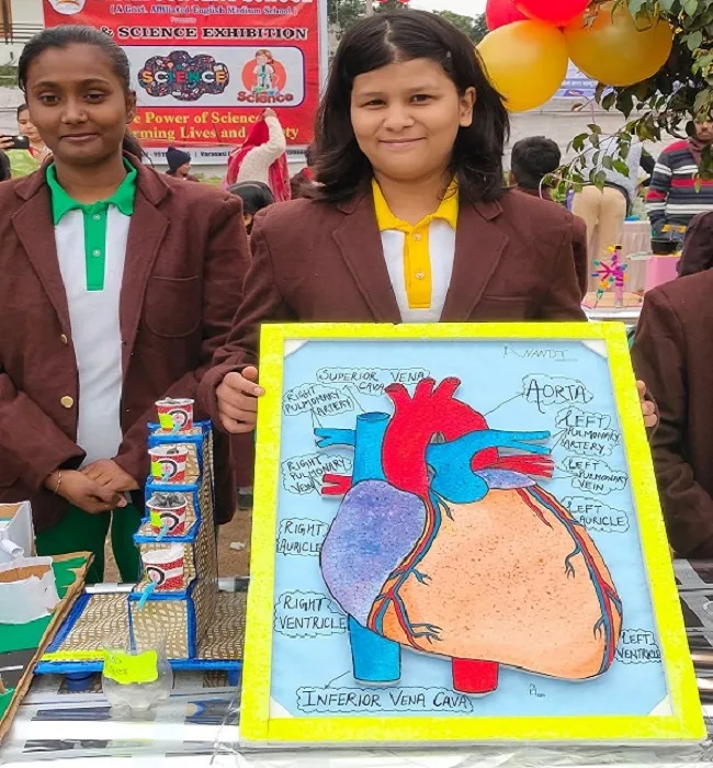 Two students in blazers present a labeled heart model at a science exhibition, with a banner, balloons, and exhibits in the background.