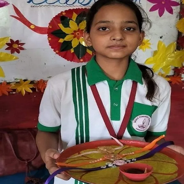 A young girl in a school uniform holds a decorated plate with rakhis and a small diya, standing in front of a colorful backdrop.