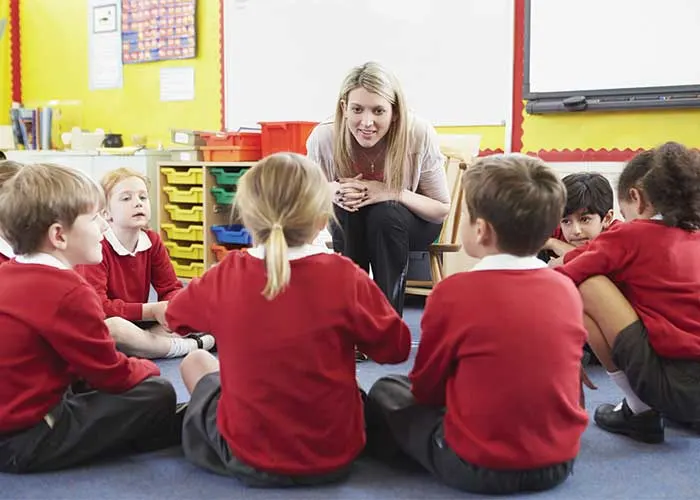 A teacher interacts with young students sitting in a circle in a bright classroom with colorful decorations.