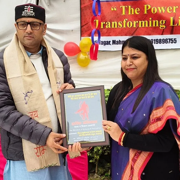 Two people holding an award plaque at an outdoor event with balloons and a banner in the background reading 'Transforming Lives'