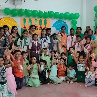 Large group of children and teachers in colorful outfits celebrating a themed event indoors, with green balloon decorations and posters