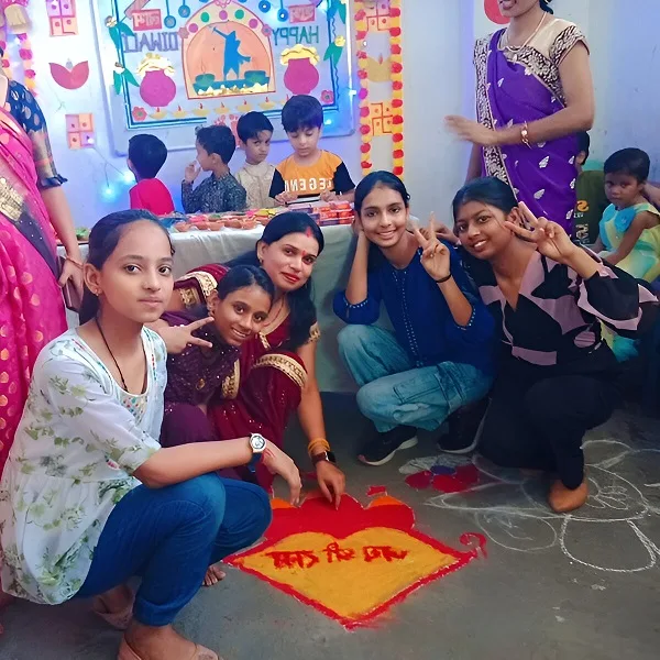 Group of girls and women posing around a colorful rangoli during a Diwali celebration with children and decorations in the background
