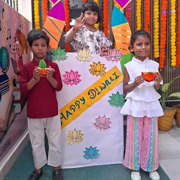 Three children posing in front of a 'Happy Diwali' board, holding handmade crafts, with festive decorations in the background