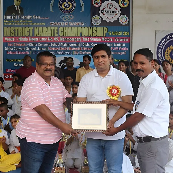 Three men at a District Karate Championship event, one receiving a framed certificate while others look on, surrounded by spectators